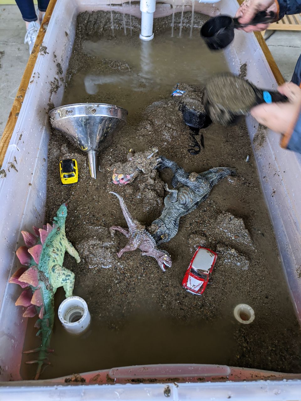 the mud table filled with various dinosaurs, toy cars, and kitchen implements